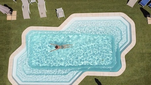Vista aérea de una piscina con una persona nadando, rodeada de sillas de descanso y sombrillas.