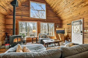 A cozy cabin interior with a fireplace, a sofa, a TV, and large windows. - Serenity Woods - Fireside Relaxation, Pet-Friendly Fun & Scenic Gazebo Views (Blue Ridge)