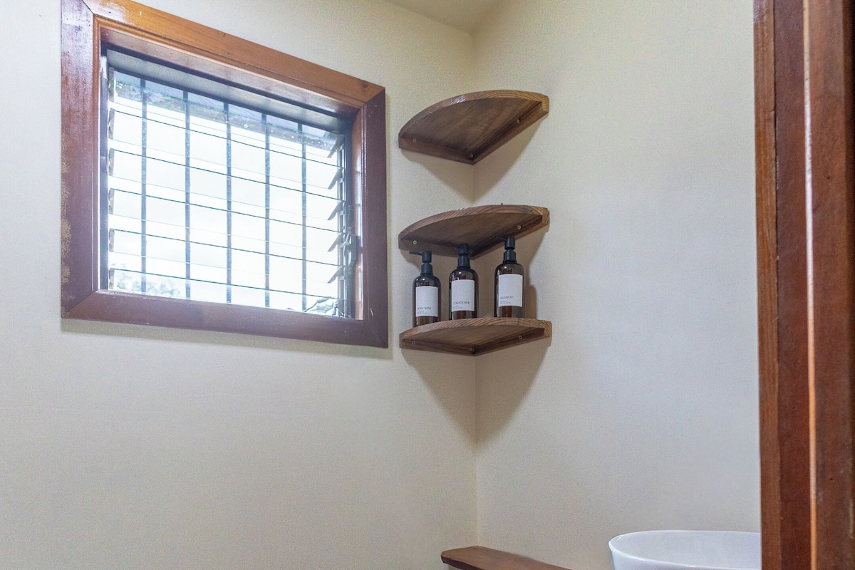 A small bathroom with a toilet, a window, and wooden shelves with bottles.