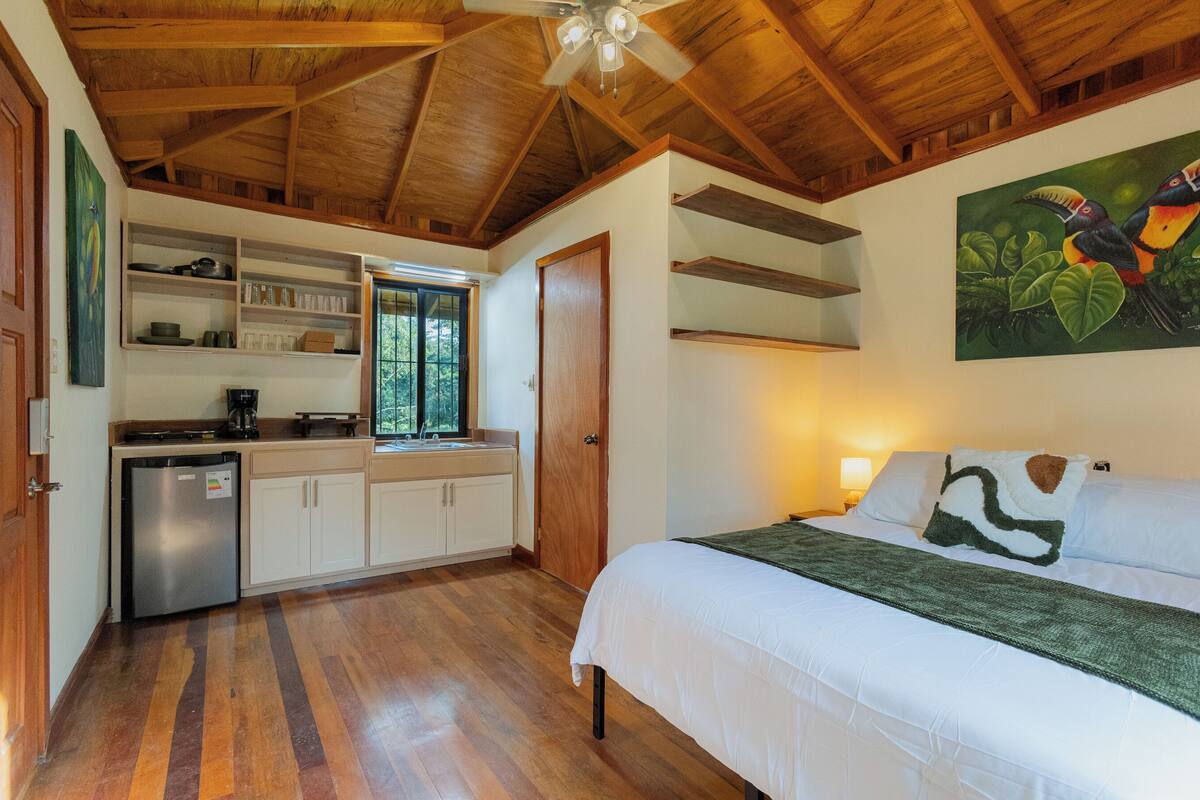 A bedroom with a bed, a nightstand, a painting, and a kitchenette with a refrigerator.