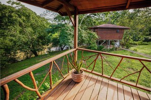 A wooden deck with a railing, a potted plant, and a view of a house in a lush green landscape. - Tohja' Riverside Cabins (San Ignacio)