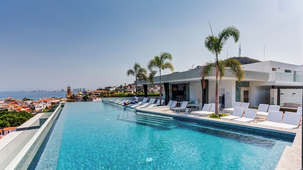 Ein Pool mit Liegestühlen und Palmen, mit Blick auf eine Küstenstadt.