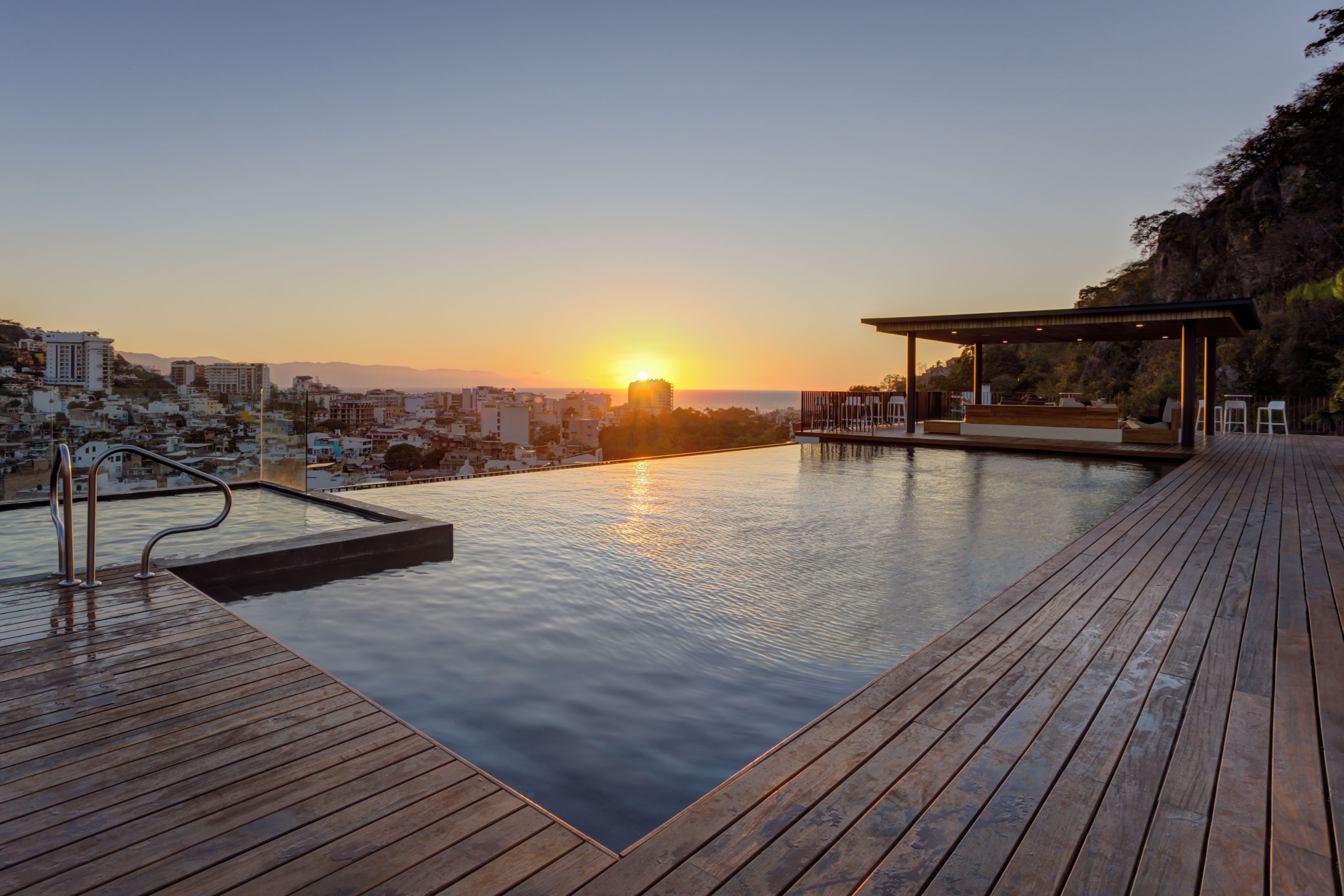 Une piscine à débordement avec une terrasse en bois et une vue sur le paysage urbain au coucher du soleil.