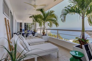 Ein Balkon mit weißen Liegestühlen, einem Deckenventilator und Blick auf das Meer.