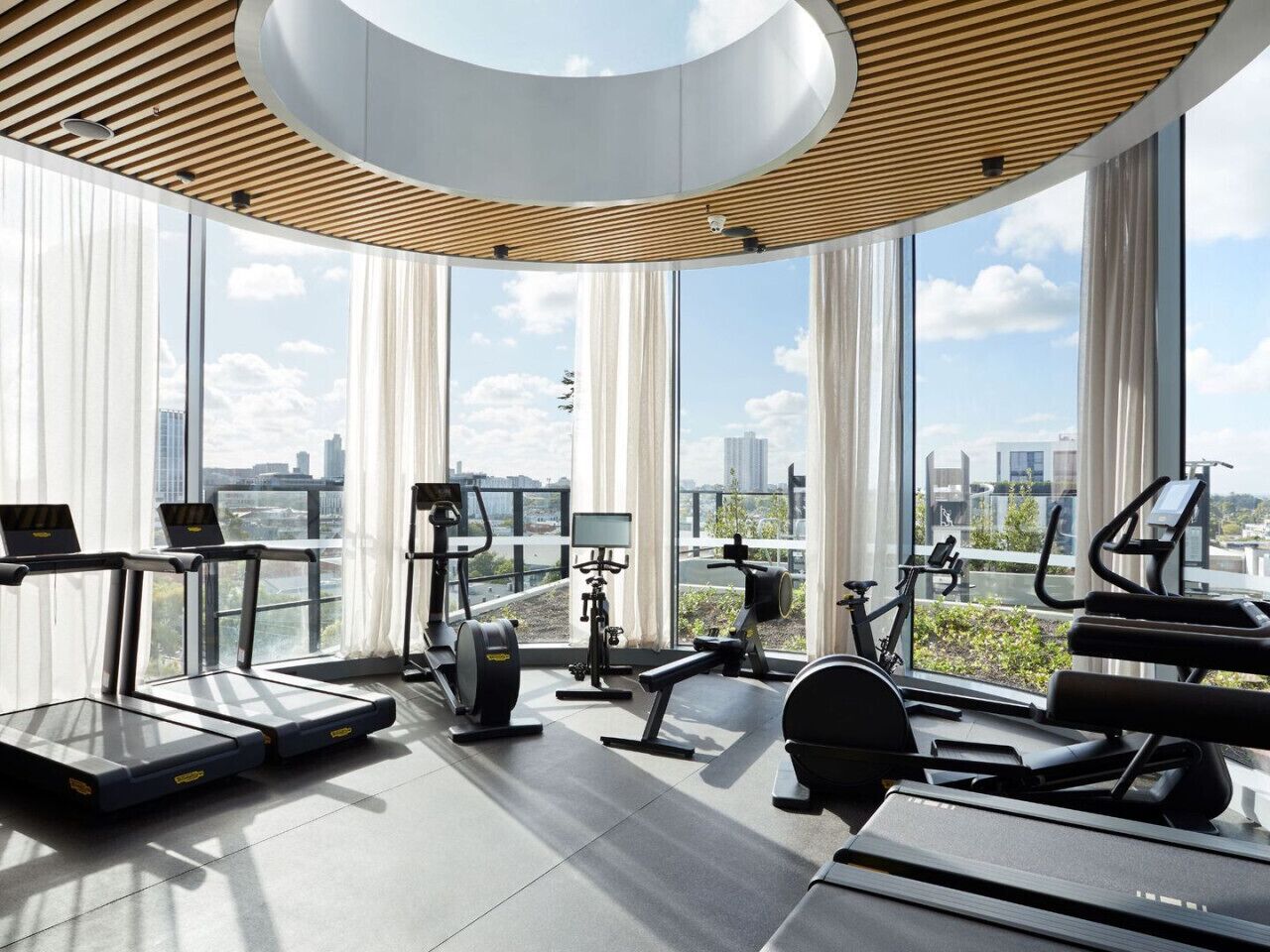 Ein modernes Fitnessstudio mit Laufbändern, Crosstrainern und Krafttrainingsgeräten, große Fenster mit Blick auf die Stadt.