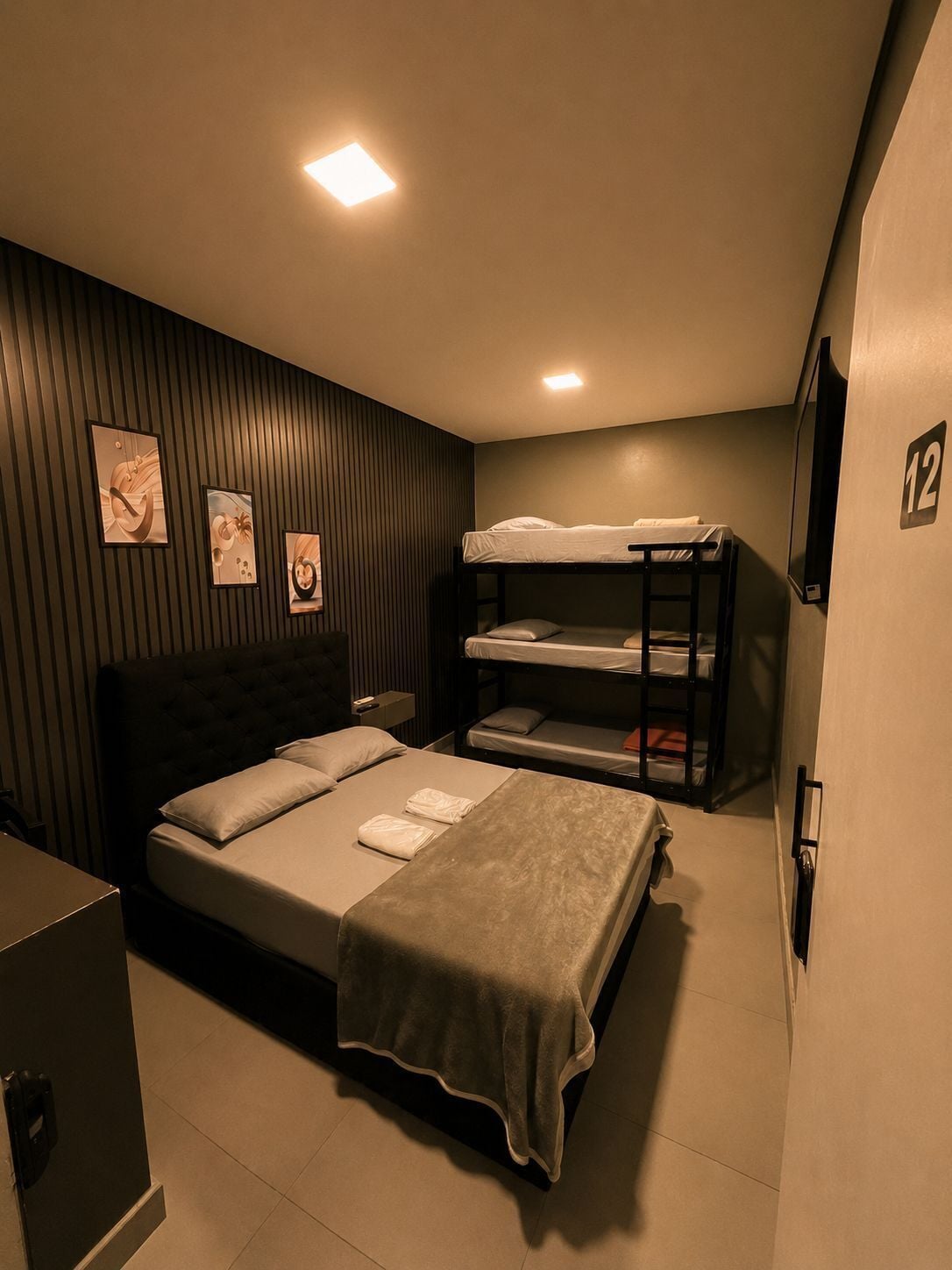 A hotel room with a bed, bunk beds, a bedside table, a wall-mounted TV, and framed artwork.
