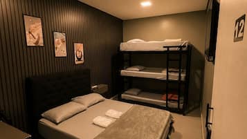 Habitación de hotel con cama, literas, mesita de noche, televisor montado en la pared y cuadros enmarcados.