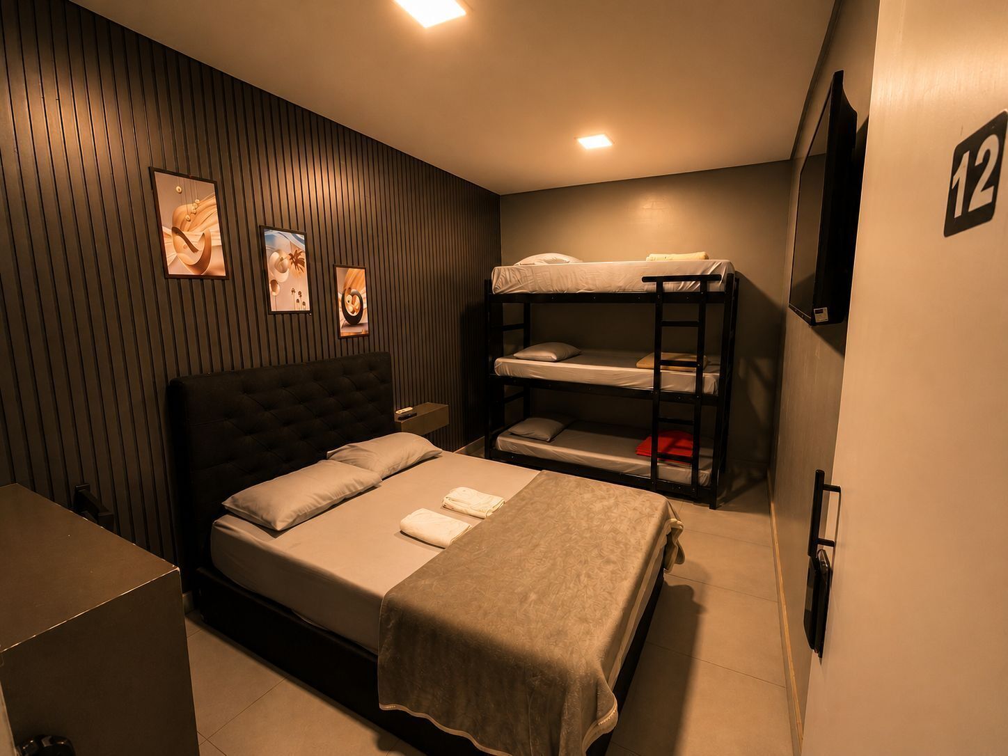 A hotel room with a single bed, a bunk bed, a flat-screen TV, and a wall-mounted number '12'.