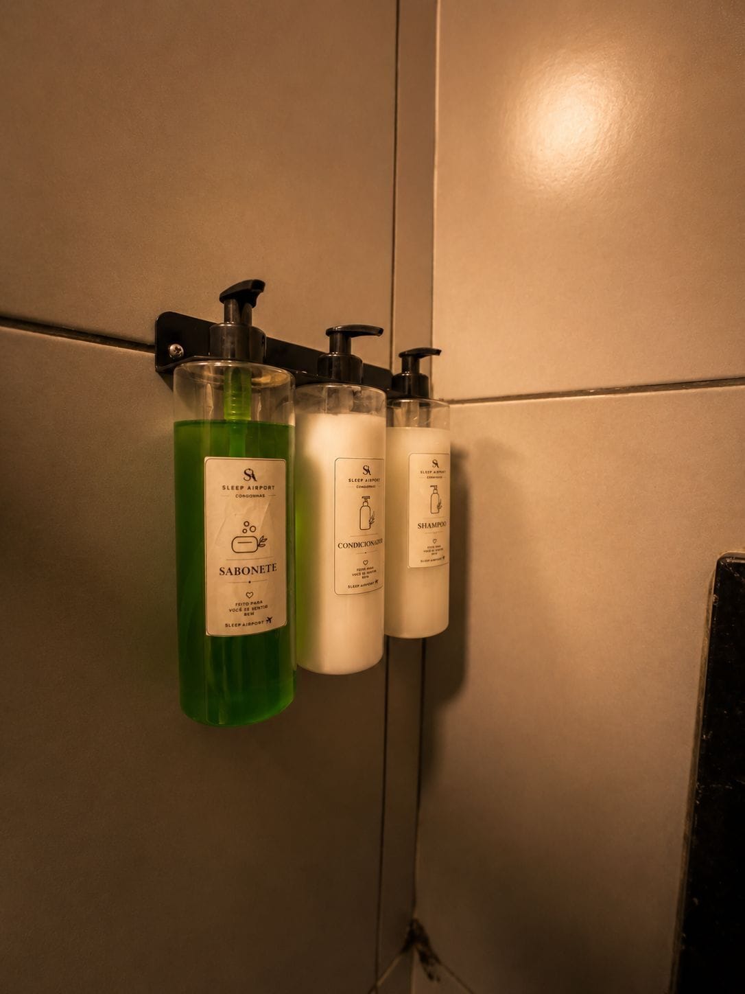 Drei Pflegeprodukte an der Wand: eine grüne Flüssigseife, ein weißes Conditioner und eine weiße Shampoo-Flasche.