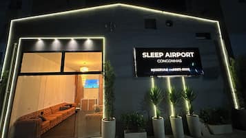 Ein Gebäudeeingang mit einem Schild, auf dem 'SLEEP AIRPORT CONGONHAS' steht, und ein Empfangsbereich mit einer Couch und einem Fernseher.