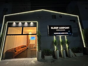 Entrada de un edificio con un cartel que dice 'SLEEP AIRPORT CONGONHAS' y una zona de recepción con un sofá y televisor.