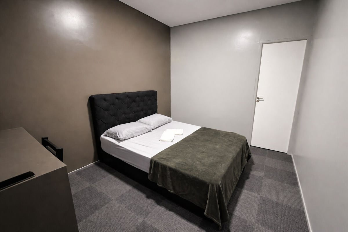 A single bed with a dark headboard, a white pillow, and a green blanket. There is a white door to the right and a small desk with a chair to the left.