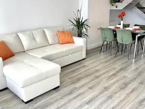 Una sala moderna con un sofá seccional blanco, sillas verdes, una mesa de comedor y una planta en maceta.