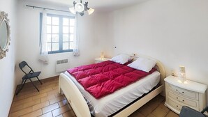 A bedroom with a bed, a chair, a dresser, a nightstand, a window, and a ceiling light fixture. - APPARTEMENT PROCHE PLAGE (Valras-Plage)