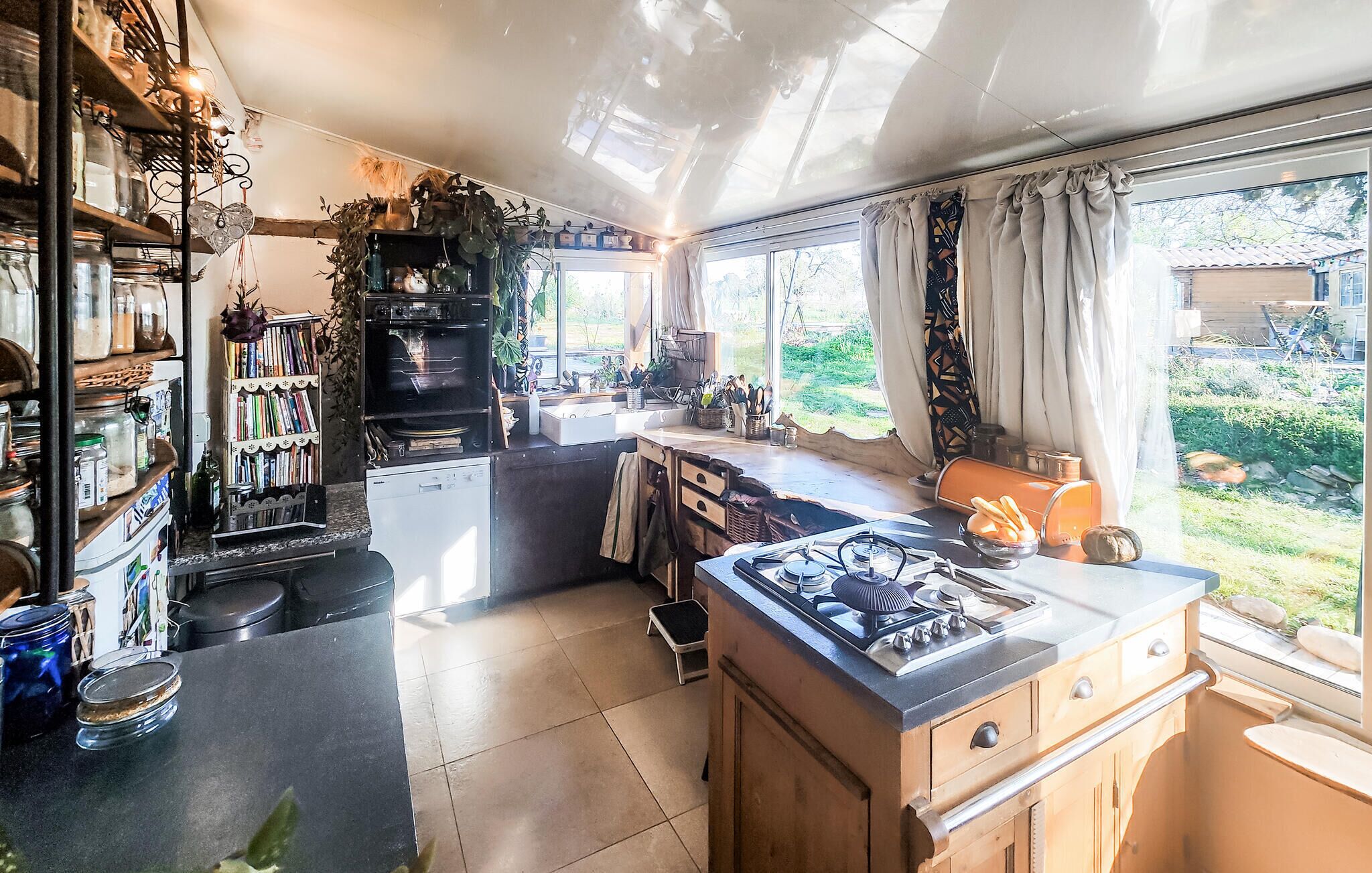 Une cuisine équipée d’un four encastré, d’une cuisinière à gaz et d’un évier. On y trouve une fenêtre avec des rideaux, ainsi qu’une bibliothèque remplie de livres et d’objets décoratifs.