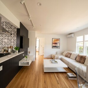 Eine moderne Küche mit dunklen Schränken, einer weißen Arbeitsplatte und Holzboden. Ein Essbereich mit weißem Tisch und einer Couch mit Kissen.