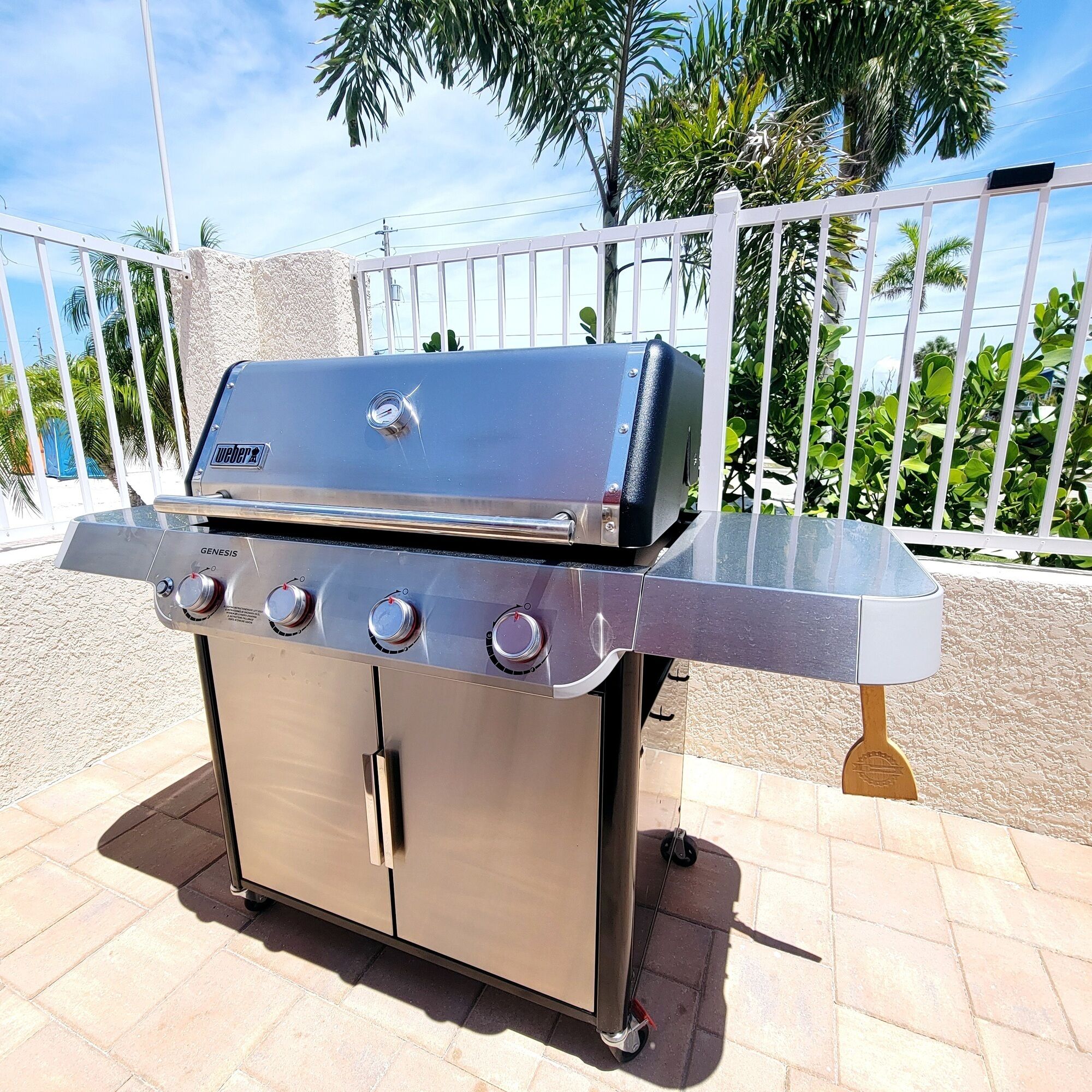 Ein Grill aus Edelstahl mit vier Brennern und einer seitlichen Ablagefläche, platziert auf einer Terrasse mit weißem Zaun und Palmen im Hintergrund.