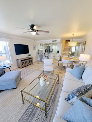 A modern living room with a sofa, coffee table, TV, and a kitchen area in the background. - BEACH FRONT PENTHOUSE w/ ROOFTOP DECK! - {Dominica 2} New 2 BR / 2 BA - Walk to Times Square! (Fort Myers Beach)