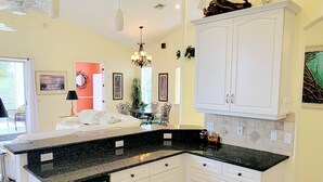 A kitchen with white cabinets, black countertops, and a built-in oven. There is a dining area with a table and chairs, and a window with curtains. - !Fun for Families! Large Pool Home + Game Room! (Fort Myers Beach)