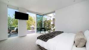 A modern bedroom with a large bed, a flat-screen TV mounted on the wall, and a sliding glass door leading to an outdoor patio. - The Art House by the Beach (Brighton)