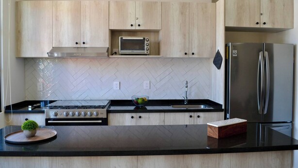 A modern kitchen with wooden cabinets, a microwave, a stainless steel refrigerator, a gas stove, and a herringbone-patterned backsplash. - Encanto Department We Bill (San Miguel de Allende)