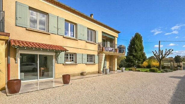 Un edificio su due piani con un balcone, una strada di accesso sterrata e un grande albero sullo sfondo.