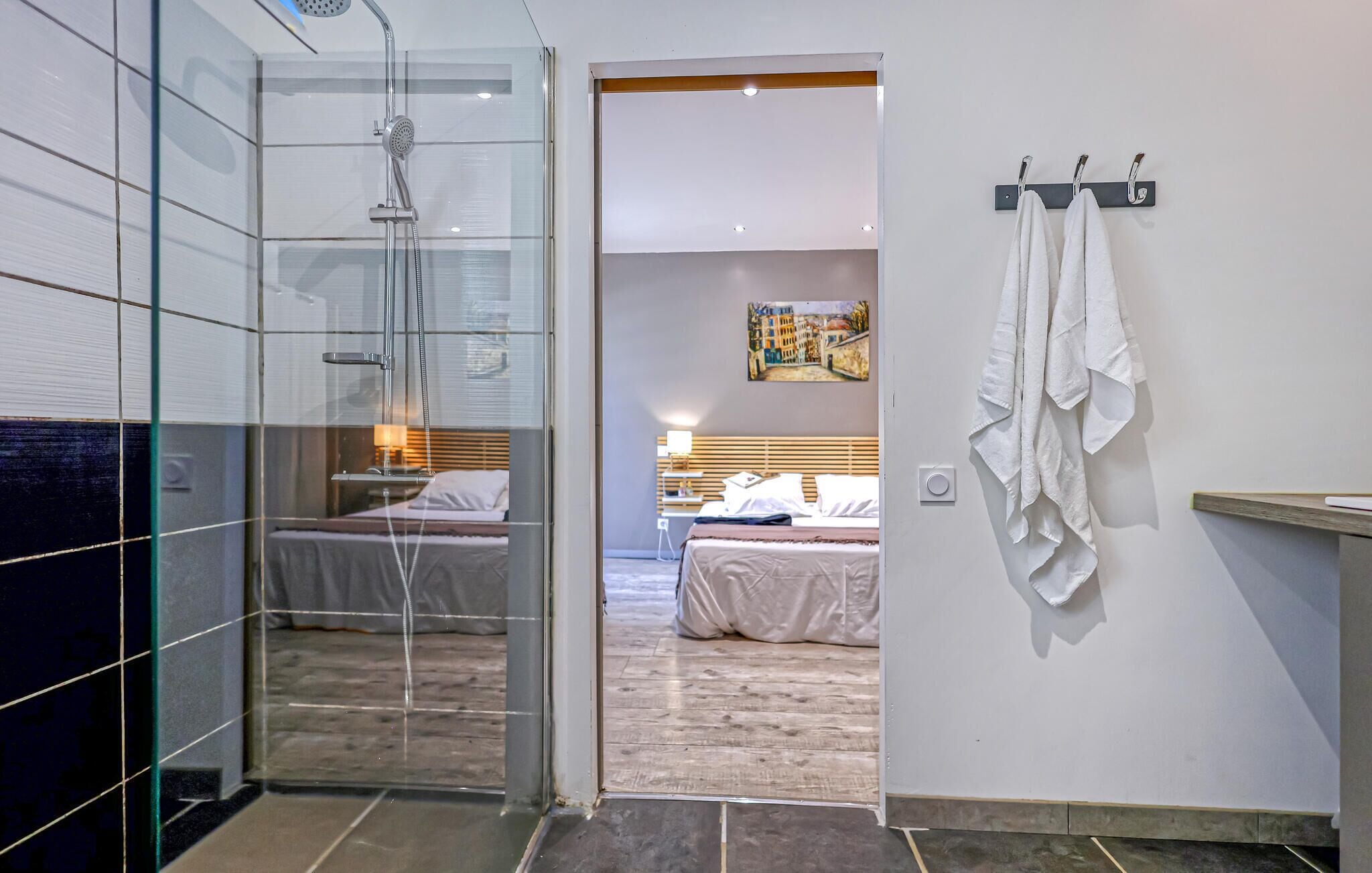 A modern bathroom with a glass shower enclosure, a visible bed with white bedding, and a towel rack with towels.
