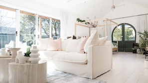 A spacious living room with a white sofa, a wooden coffee table, and a large window providing a view of the outdoors.