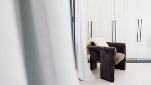 Een kamer met een donkerbruine stoel, witte gordijnen en witte kasten.