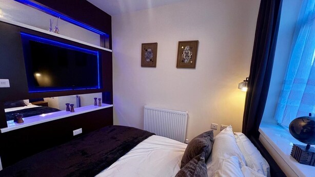A hotel room with a bed, a flat-screen TV, two wall-mounted artworks, a window with curtains, and a radiator. - Beautiful 2-bedroom apartment near LHR (Feltham)