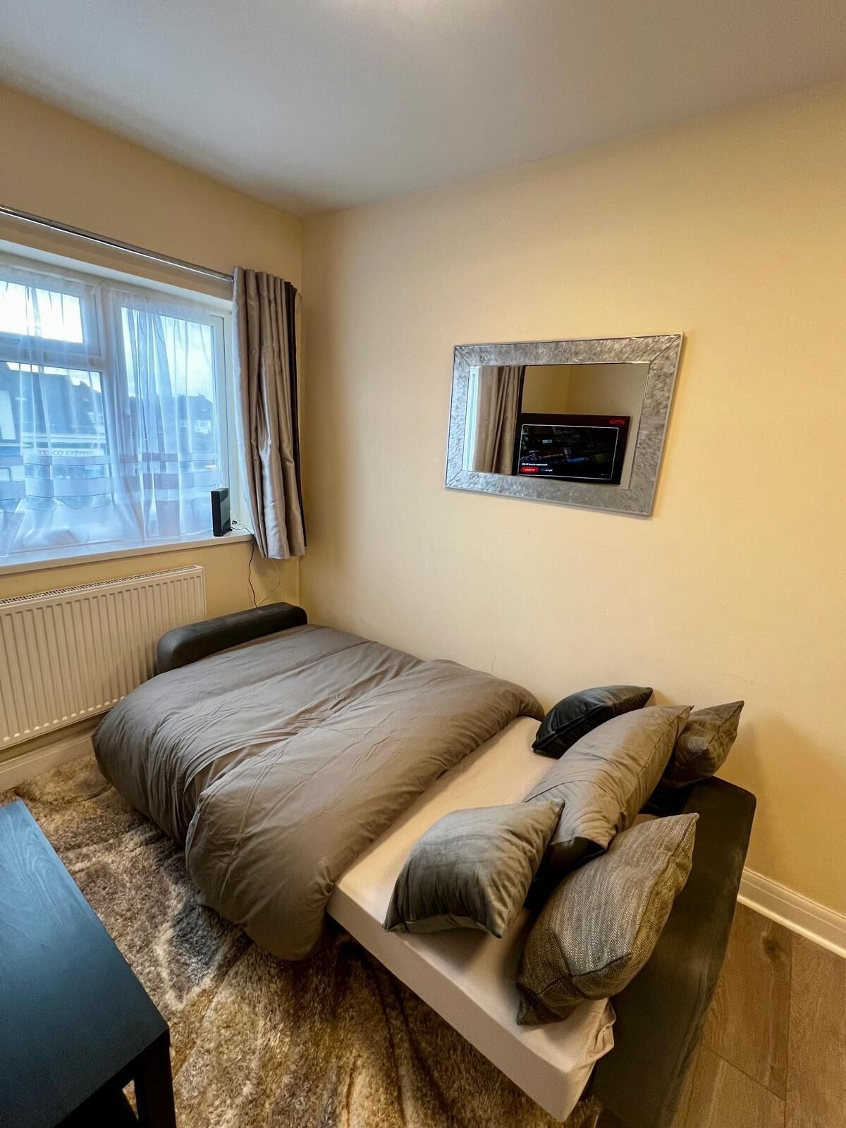 A bedroom with a bed, a bedside table, a mirror, and a window with curtains.