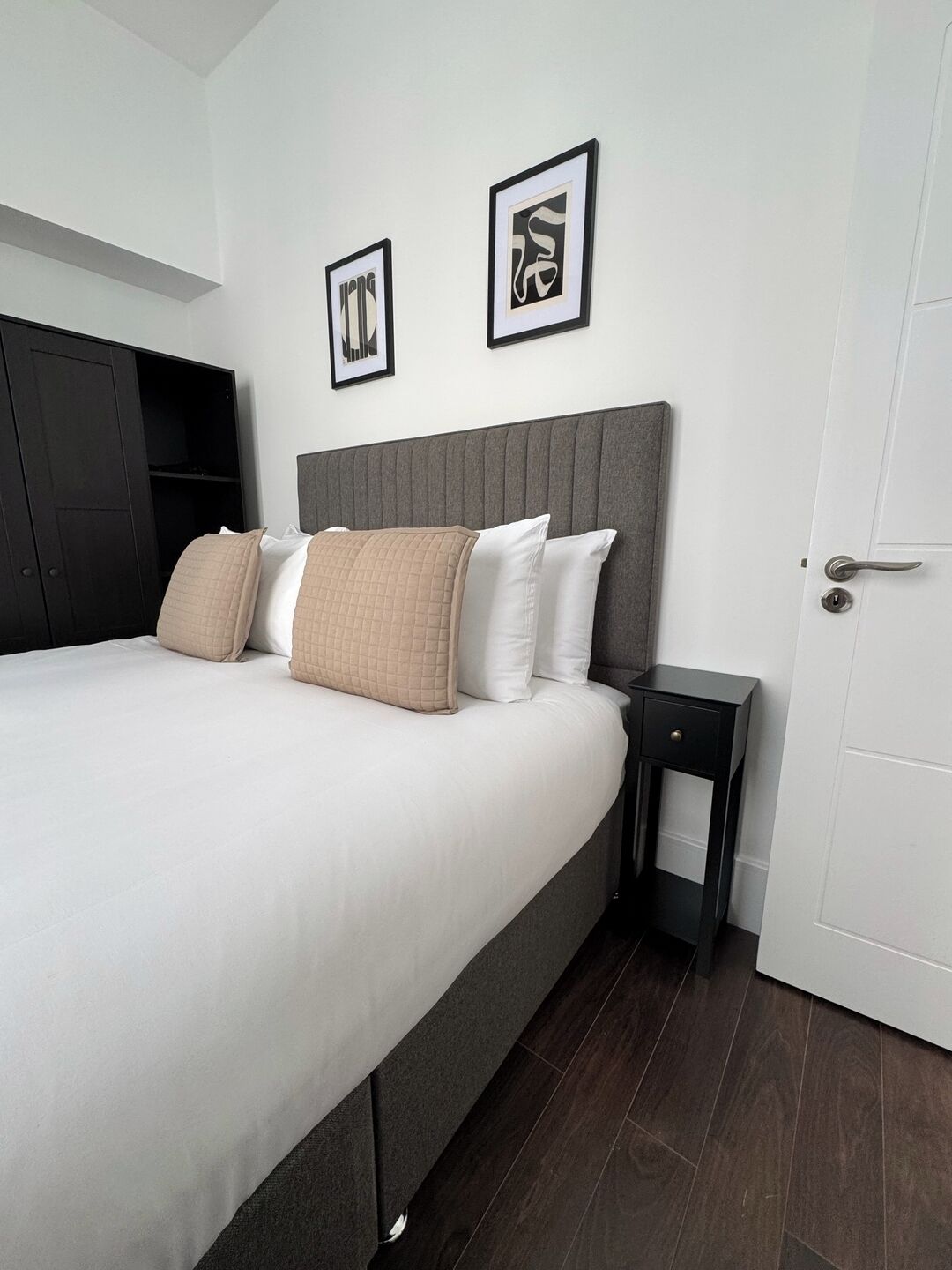 Una cama bien hecha con cabecera gris, una mesita de noche y dos cuadros en la pared.