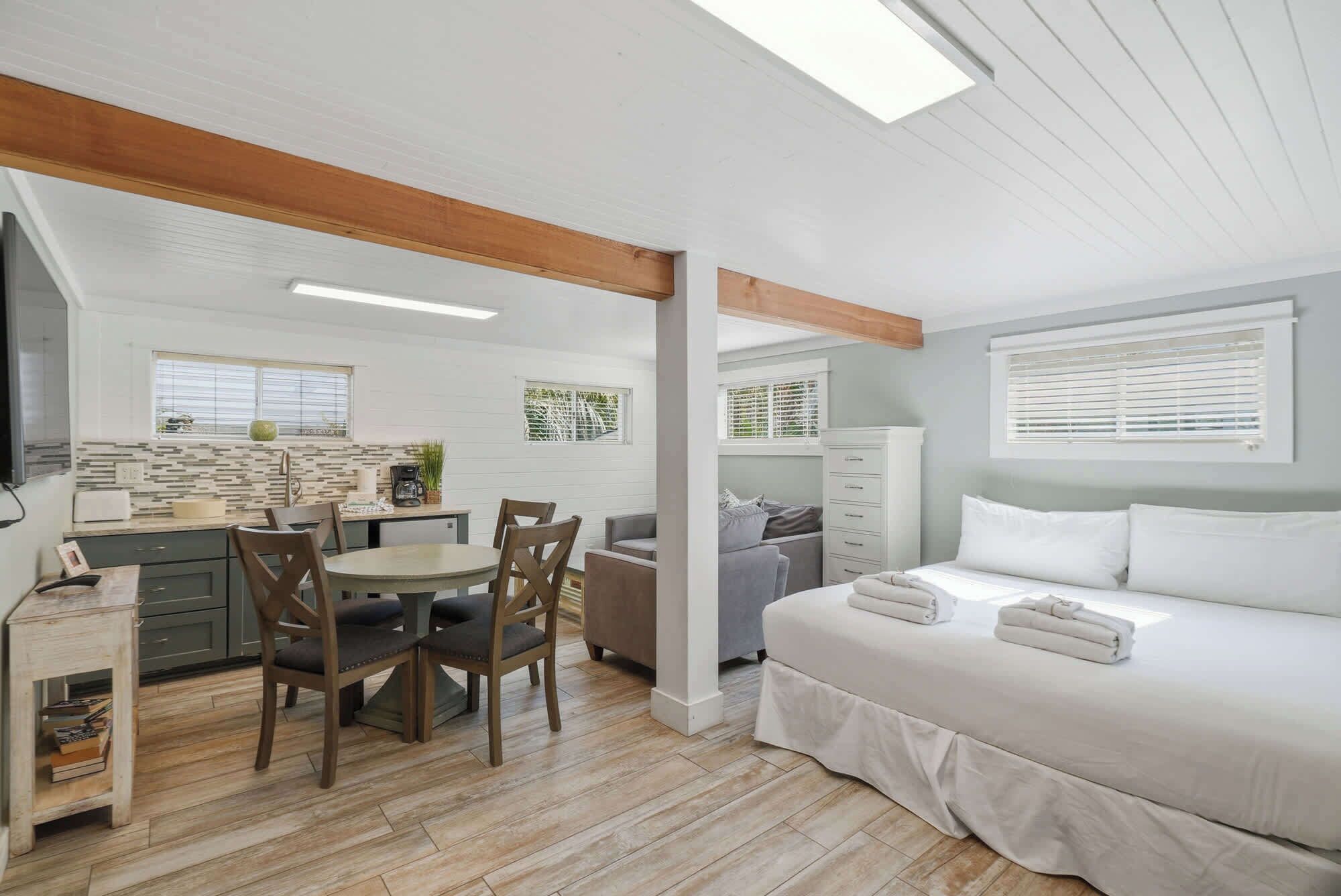 A room with a bed, a dining table with chairs, a kitchenette, and a window with blinds.