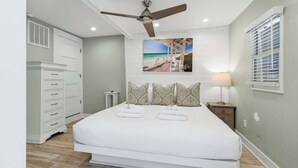 Ein Schlafzimmer mit Bett, Deckenventilator, Nachttisch mit Lampe, Kommode und Fenster mit Jalousien.