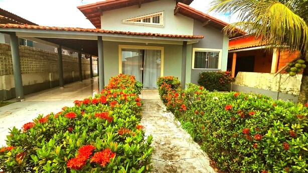 A two-story building with a tiled roof, a covered entrance, and a pathway lined with red flowers and green shrubs. - SaltBeach Três Quartos no Atalaia (Salinópolis)