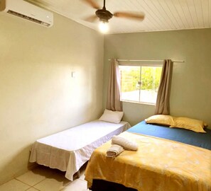 Acomodação simples, com um quarto único, duas camas, ventoinha no teto, ar condicionado e janela com cortinas.