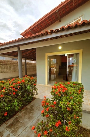 Uma área externa coberta com um alpendre, uma porta de vidro deslizante que dá para um espaço interno, e dois arbustos floridos com flores vermelhas.