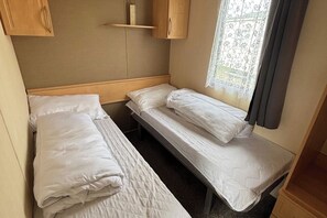 A small, compact room with two single beds, a wooden headboard, a shelf above the beds, and a window with curtains. - Seaside Tranquility Retreat in Clacton (Clacton-on-Sea)