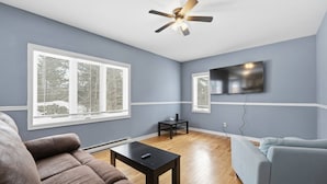 A living room with a ceiling fan, a flat-screen TV, a sofa, a coffee table, and large windows. - Executive 7 Bedrooms 3 5 Bathrooms (Bedford)