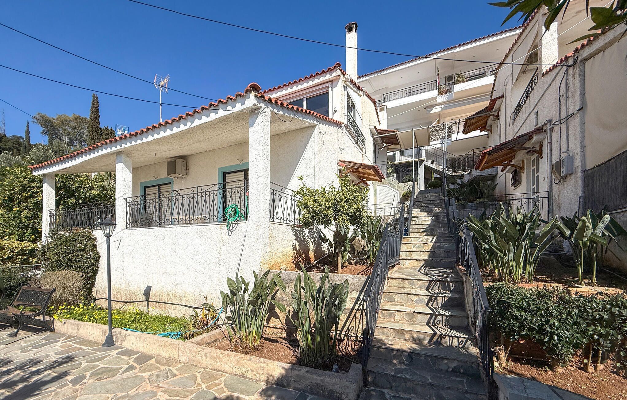 Un bâtiment à deux étages doté d’un escalier en pierre, d’un balcon avec des rampes en métal et d’un jardin où poussent différentes espèces de plantes.