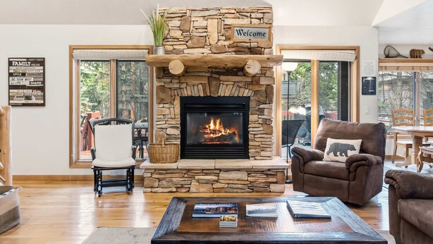 Una chimenea de piedra con un cartel que dice 'Welcome', una silla blanca, un sillón marrón con un cojín de oso, y una mesa con libros y una cesta.
