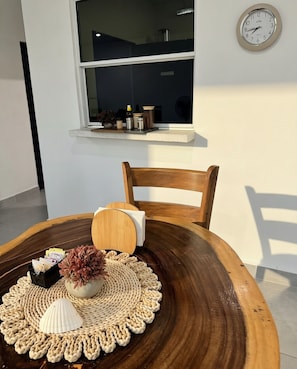 Une table ronde en bois, sur laquelle repose un set de table en fibres tressées, un vase contenant des fleurs séchées et un petit récipient rempli de monnaie. Une chaise en bois est placée derrière la table.