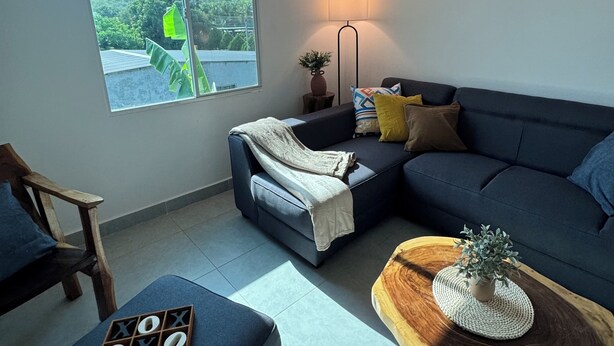 A modern living room with a sectional sofa, a small table with a plant, a lamp, and a window with a view of greenery. - Surf City Private 7 Suite Hotel with AC + hot water (Conchalio)