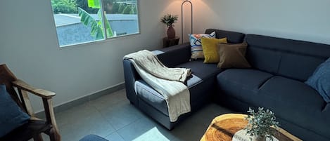 A modern living room with a sectional sofa, a small table with a plant, a lamp, and a window with a view of greenery.