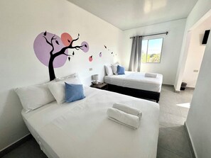 Une chambre d’hôtel avec deux lits, une fresque murale, une fenêtre avec des rideaux et une petite table avec une lampe.