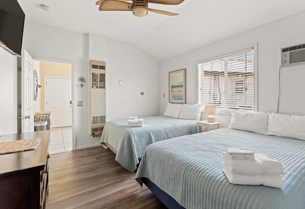 A bedroom with two beds, a ceiling fan, a window with blinds, and a bedside table with a lamp.