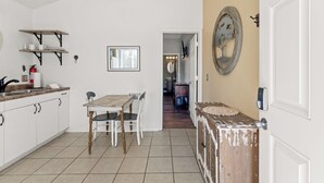 Een keuken met witte kasten, een eettafel met stoelen, een houten kast met een decoratieve spiegel, en een deur met een lichtschakelaar.