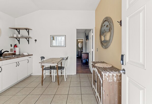 A kitchen with white cabinets, a dining table with chairs, a wooden cabinet with a decorative mirror, and a door with a light switch. - Snug Harbor Inn | The beach suite | Near Lake Michigan (Muskegon)