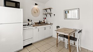 Een keuken met een witte koelkast, fornuis en magnetron. Er is een houten tafel met twee stoelen en een wandplank boven de gootsteen.
