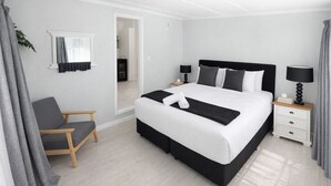 A modern hotel room with a large bed, a bedside table with a lamp, a chair, and a view through an open door.